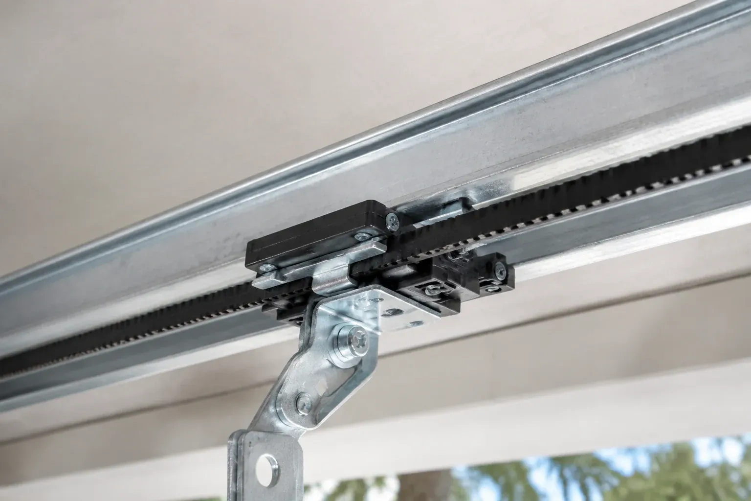 Black plastic trolley and bent galvanized bracket on a steel opener rail with a toothed black belt, against a ceiling. — Belt-Drive Opener Repair, Mississauga